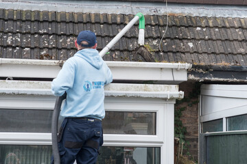 A window and gutter cleaner cleaning Dirty clogged white plastic pvc gutters and drain pipes with mossy green mould on plastic fascias. Blocked drains and guttering need  regular yard work maintenance