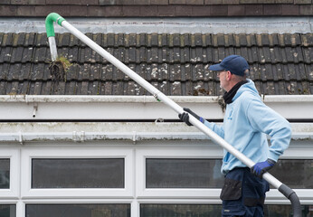 A window and gutter cleaner cleaning Dirty clogged white plastic pvc gutters and drain pipes with mossy green mould on plastic fascias. Blocked drains and guttering need  regular yard work maintenance