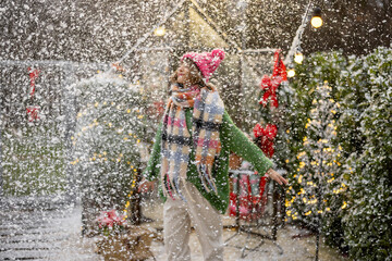 Woman enjoys the snowfall while dancing at beautifully decorated backyard. Background image, person is out of focus. Concept of winter holidays