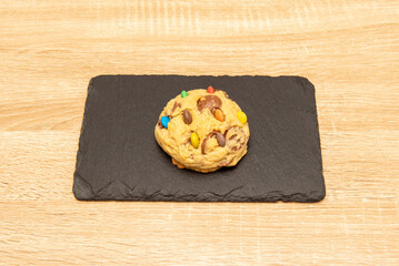 Delicious fluffy cookie with colored chocolate chips on a black slate plate on an oak table
