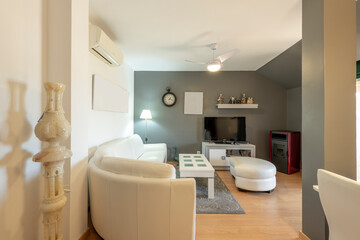 Living room of a house with a white leather sofa covering one wall and a dining table and a matching coffee table, flat tv on a sideboard, a column and a gray painted wall