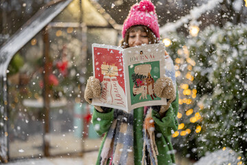 Young woman reads some magazine on New Year's theme at snowy backyard decorated for a winter...