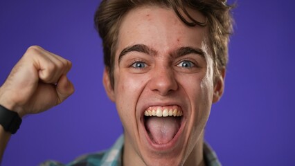 Closeup of excited jubilant overjoyed young man 20s doing winner gesture celebrate clenched fist say yes isolated on purple background studio portrait