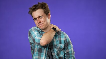 Portrait of young hipster man 20s with sore neck shoulder problem isolated on purple background in studio