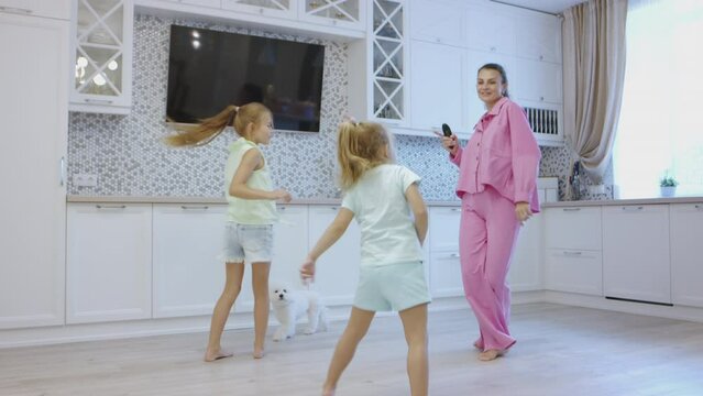 Happy mother enjoys dancing with daughters and fluffy doggy in kitchen with stylish interior and TV screen on wall. Young woman in pink pajamas and little girls spend weekend at home together