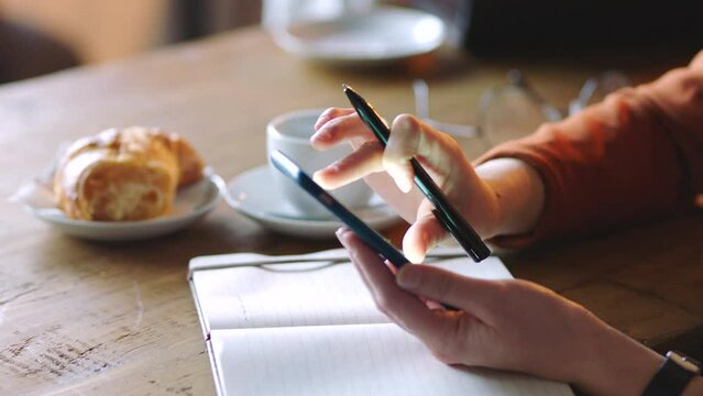 Coffee shop, smartphone and hands with notebook for creative inspiration, social media marketing strategy and remote work planning. Croissant, tea and cafe table and digital writer with writing ideas