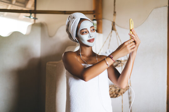 Young Asian female shooting beauty vlog while doing mask procedures for nourishing treatment in home bathing using front camera on digital cellular, attractive woman in earphones making selfie photos