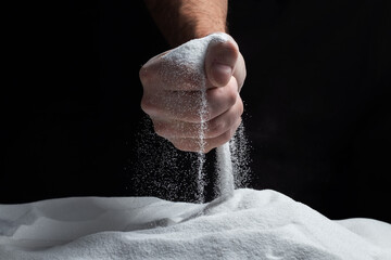 Man with handful of white dry sand in her hands, spilling sand through fingers on black background.