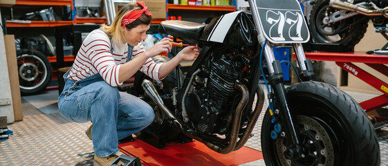Mechanic woman with screwdriver repairing custom motorcycle over platform on factory