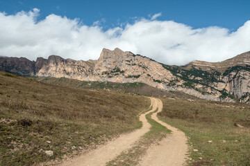 Old path through mountains. Trekking mountain trail. Bright atmospheric minimalist alpine landscape with stony footpath among grasses in highlands. Pathway uphill. Way up mountainside.