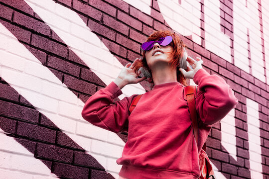 Stylish woman in magenta color jacket and heart shaped sunglasses wearing wireless headphones on her head and listening music. Urban city street fashion. Color of the 2023 year. Selective focus.