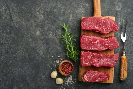 Raw steaks. Top blade steaks on wood burning board with spices, rosemary, vegetables and ingredients for cooking on black background. Top view.  Copy space.