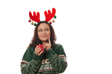 Isolated on white background, charming delightful multi-ethnic woman in warm Xmas patterned green sweater and deer antler hoop, holding a red lit Christmas candle and smiling looking up at copy space