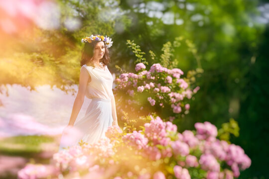 Outdoor portrait of young happy smiling fantasy woman walking among flowers. Young beautiful goddess enjoys spring nature in bright sunlight. Fairy tale image art photo with pink roses frame