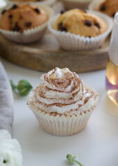 Muffins with cream and chocolate sprinkles on a wooden stand near white ranunculus and a cup of tea