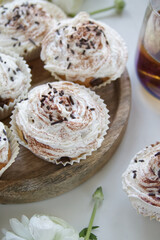 Muffins with cream and chocolate sprinkles on a wooden stand near white ranunculus and a cup of tea