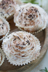 Muffins with cream and chocolate sprinkles on a wooden stand near white ranunculus and a cup of tea