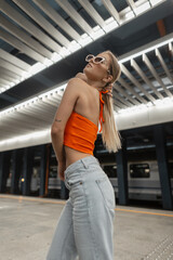 Cool urban fashion woman model with cool sunglasses in trendy casual wear with orange top and vintage jeans in the subway