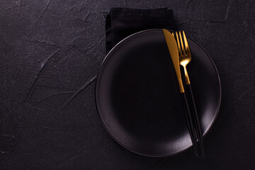 Modern table setting. Black plate, knife, fork and grey napkin on textured  black background. Top...