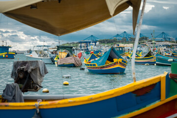 Luzzu, typical fishing boats of Malta moored in the marina of Marsaxlokk on an autumn day. Many ships in the bay, container cranes in the background.