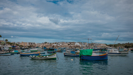 Fototapeta premium Luzzu, typical fishing boats of Malta moored in the marina of Marsaxlokk on an autumn day. Many ships in the bay, cityscape in the background.
