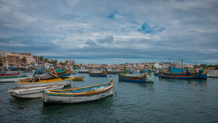 Fototapeta na wymiar Luzzu, typical fishing boats of Malta moored in the marina of Marsaxlokk on an autumn day. Many ships in the bay, cityscape in the background.