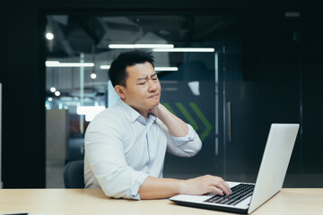 Plakat Office work syndrome. A young Asian man sits in the office at the desk, holds his neck, feels pain, tension. He closed his eyes, massages with his hand.
