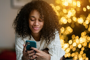 Merry Christmas and Happy Holidays! Young african american woman chatting on a smartphone on by the Christmas tree in the cozy living room of the house   