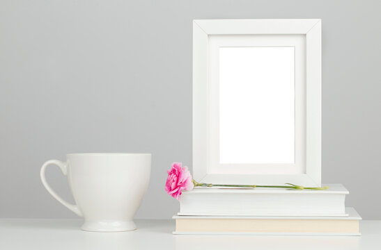White picture frame mockup on desk with books, porcelain mug and pink flower 