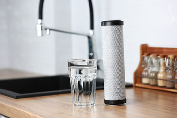 A glass of clean fresh water and carbon filter cartridge on wooden table in a kitchen interior. Installation of reverse osmosis water purification system. Concept Household filtration system.