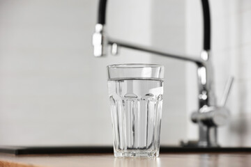 Transparent glass of clean filtering water on wooden table in kitchen interior. Tap with purified...