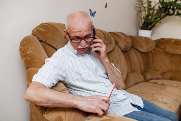 Fototapeta premium An elderly man talks on the phone and writes down the data in a diary or glider. Organization of business in retirement. A pensioner works remotely, conducts business from home