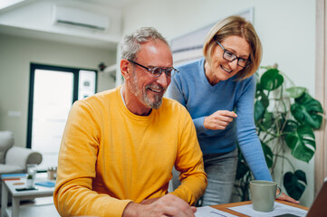 Senior middle aged happy couple using laptop together at home. Smiling elderly family reading news or shopping online. Older people and technology concept. Focus on a man in a yellow sweater.