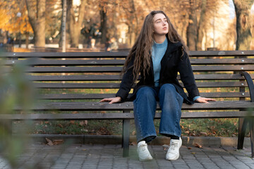 Obraz na płótnie Canvas Young woman with long brown hair sits on bench in autumn park and waits for someone. Autumn time.