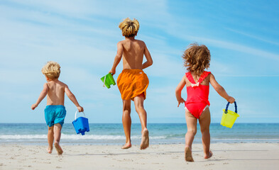 Three happy kids run on the sand beach with buckets from behind