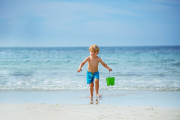 Fototapeta na wymiar Cute little blond boy run with bucket on ocean beach from waves