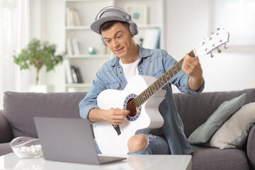 Guy with an acoustic guitar looking at a computer at home wearing headphones and playing