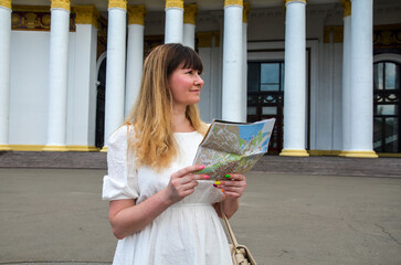 Smiling woman traveler in white dress with map at town street. Concept of travel, summer vacation, solo female tourism, adventure, trip, journey.