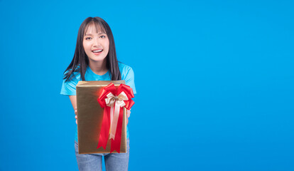 Cheerful asian woman holding gift box giving surprise family in festive eve season celebration or anniversary standing on blue background. Cute young girl give celebrate gift box gold over isolated.