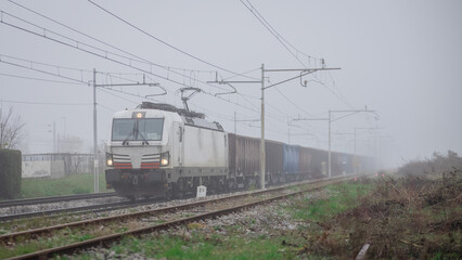 Freight train with a modern electric locomotive pulling open goods wagons or gondolas through foggy weather.