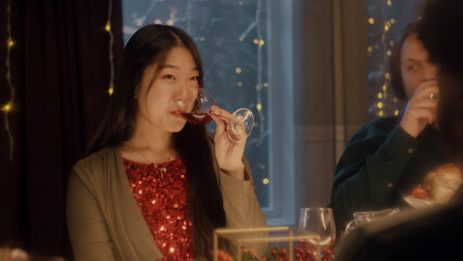 Asian woman drinks wine, talks and smile. Multi cultural friends celebrating Christmas or New Year 2023. Served holiday table. Warm atmosphere of Christmas dinner at home. Christmas party.
