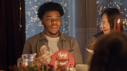 Surprised African American man opens gift. Multi cultural friends celebrating Christmas or New Year 2023. Served holiday table with different dishes. Warm atmosphere of Christmas dinner at home.