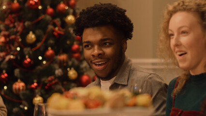 Happy multi cultural family celebrating Christmas or New Year 2023. African American man eats and smiles. Served holiday table with dishes. Warm atmosphere of family Christmas dinner at home.