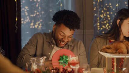 Surprised African American man opens gift. Multi cultural friends celebrating Christmas or New Year 2023. Served holiday table with different dishes. Warm atmosphere of Christmas dinner at home.