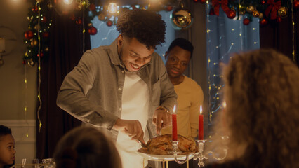 African American man cuts turkey or chicken with knife. Multi cultural friends celebrating Christmas or New Year. Served holiday table with candles. Warm atmosphere of Christmas dinner at home.