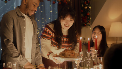 African American man cuts turkey or chicken with knife. Multi cultural friends celebrating Christmas or New Year. Served holiday table with candles. Warm atmosphere of Christmas dinner at home.