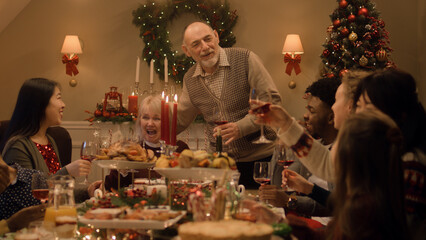 Happy multi cultural family celebrating Christmas or New Year, raising glasses with wine. Served holiday table with dishes and candles. Warm atmosphere of family Christmas dinner at home. Cheers.