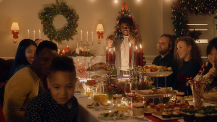 African American man givs gift to young boy. Happy family celebrating Christmas or New Year 2023. Served holiday table with dishes and candles. Warm atmosphere of family Christmas dinner at home.
