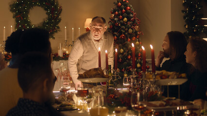 Elderly grandfather tells joke or life story to his large multi cultural family. They celebrating Christmas. Served table with dishes and candles. Warm atmosphere of family Christmas dinner at home.