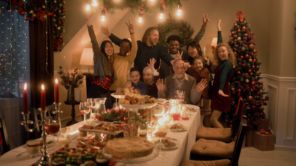 Asian woman sets timer on camera or phone. Happy multi cultural family takes group photo. They celebrating Christmas or New Year. Table with dishes and candles. Family Christmas dinner. Camera view.
